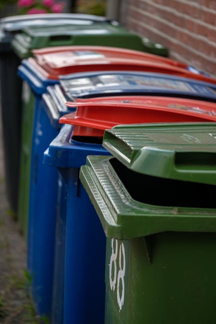 A row of outdoor bins in a residential garden setting, including green, blue, red, and black containers with closed lids, positioned alongside a brick wall. The green bin is in the foreground with a t