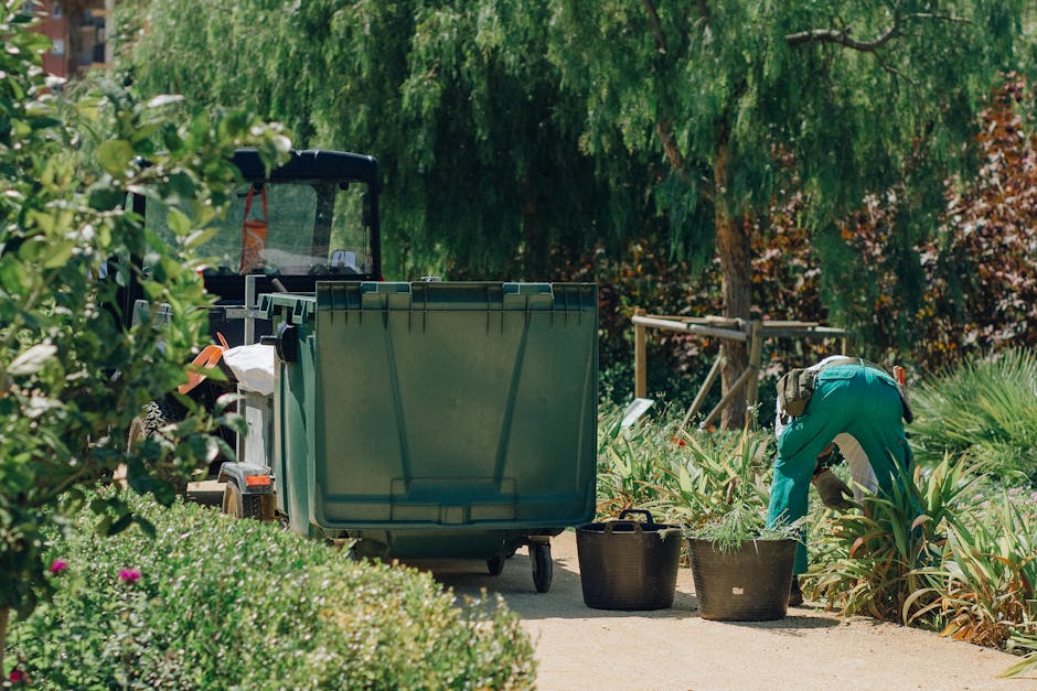 A row of outdoor bins in a residential garden setting, including green, blue, red, and black containers with closed lids, positioned alongside a brick wall. The green bin is in the foreground with a t